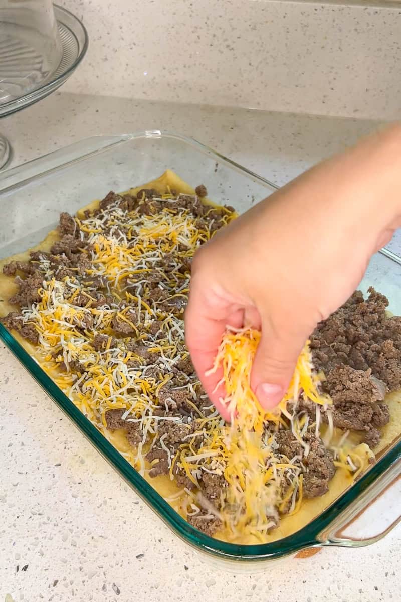 Shredded cheese being sprinkled over sausage on crescent roll dough before baking.