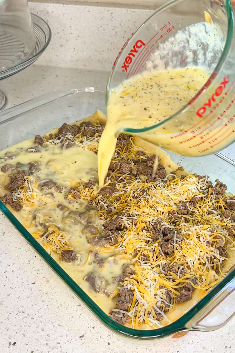 Egg mixture being poured over sausage and cheese to assemble the breakfast casserole.