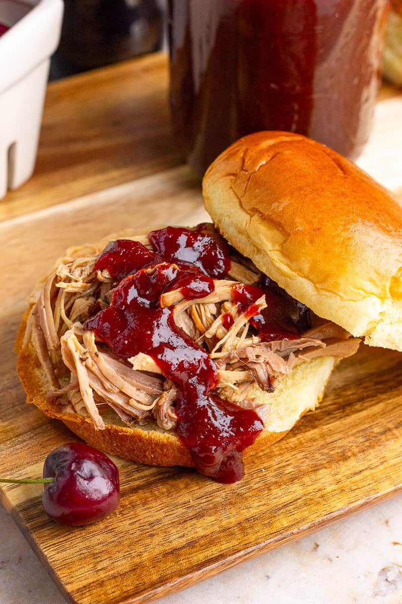 A pulled pork sandwich is topped with cherry BBQ sauce on a wooden board.