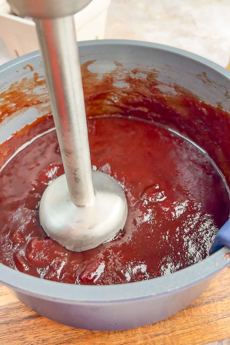 An immersion blender blends cherry cola BBQ sauce in a saucepan until smooth.