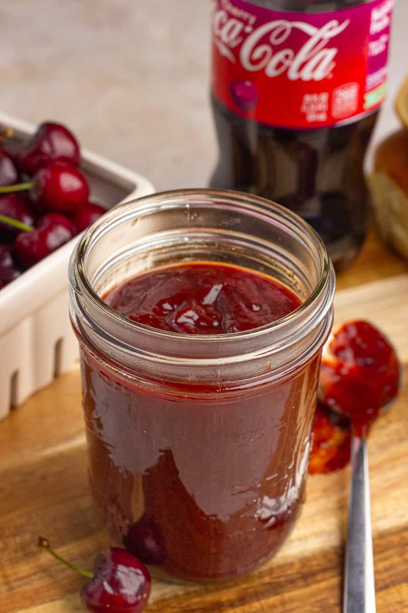 A jar of cherry cola BBQ sauce sits beside fresh cherries and a bottle of cherry cola.