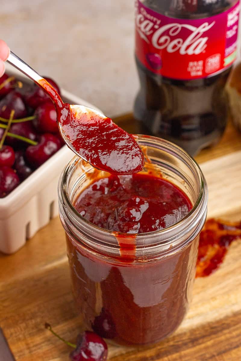 A spoon lifts thick cherry cola BBQ sauce from a jar with fresh cherries in the background.