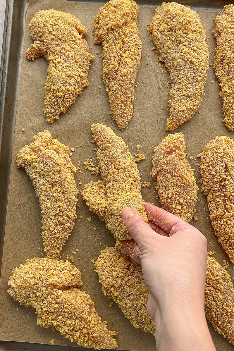 A hand places cornflake coated chicken tenders on a parchment-lined baking sheet.