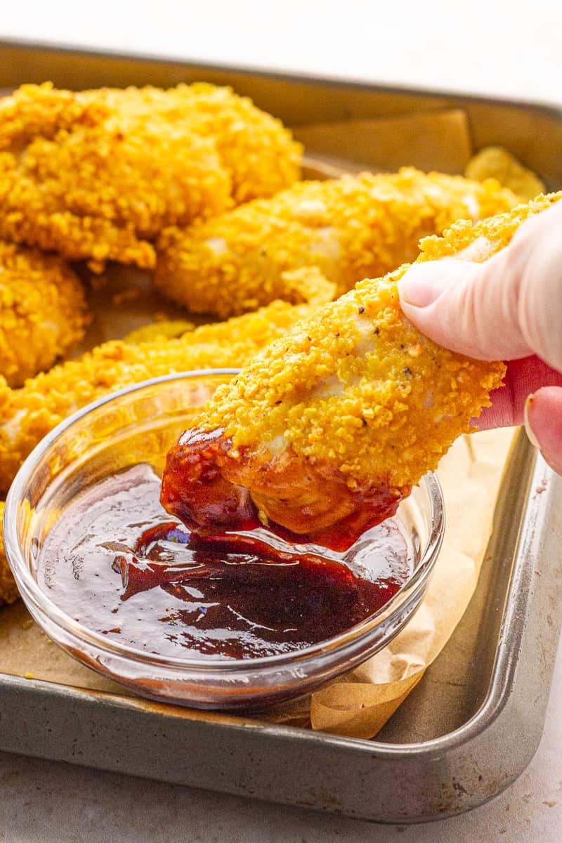 A crispy cornflake chicken tender is dipped into barbecue sauce.