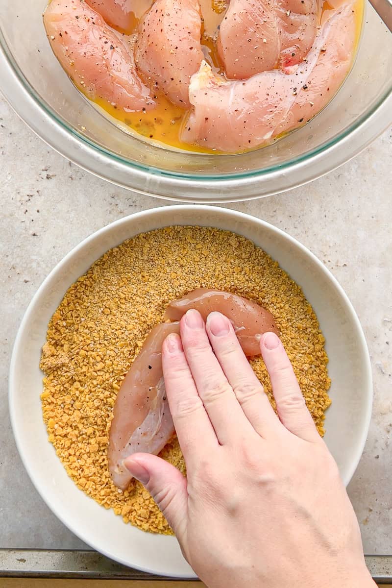 A chicken tenderloin is pressed into the seasoned cornflake coating.