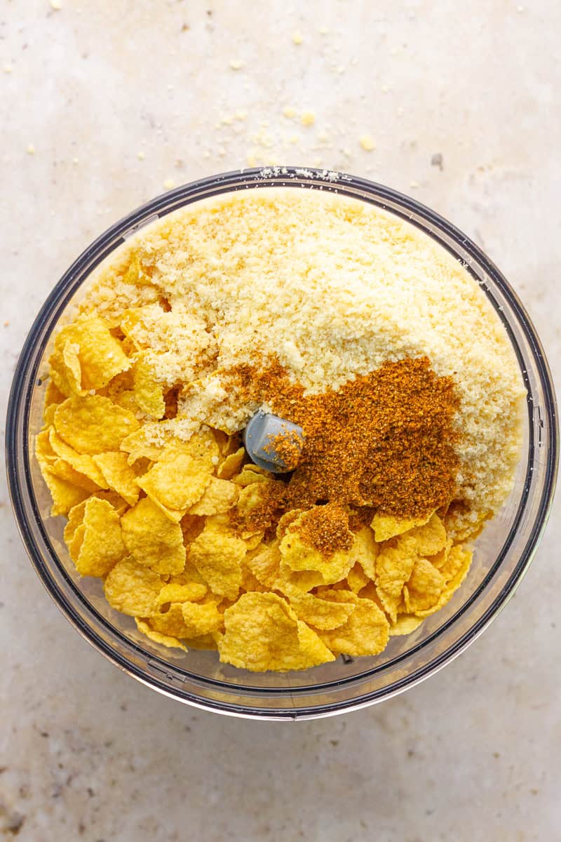 Cornflakes, grated Parmesan, and Old Bay seasoning are added to a food processor.