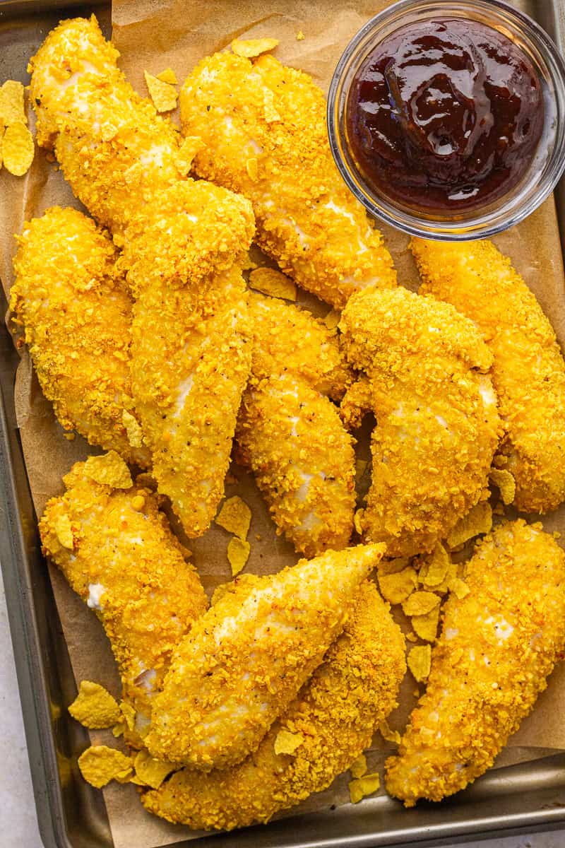Baked cornflake chicken tenders are served on a parchment-lined pan with barbecue sauce.