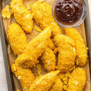 Baked cornflake chicken tenders are served on a sheet pan with barbecue sauce for dipping.