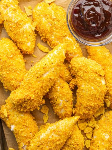 Baked cornflake chicken tenders are served on a sheet pan with barbecue sauce for dipping.