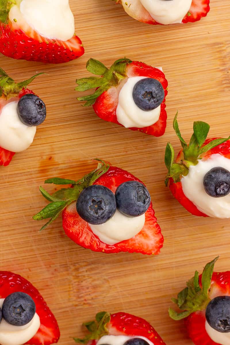 Strawberry halves filled with cheesecake filling and topped with blueberries are arranged on a cutting board.