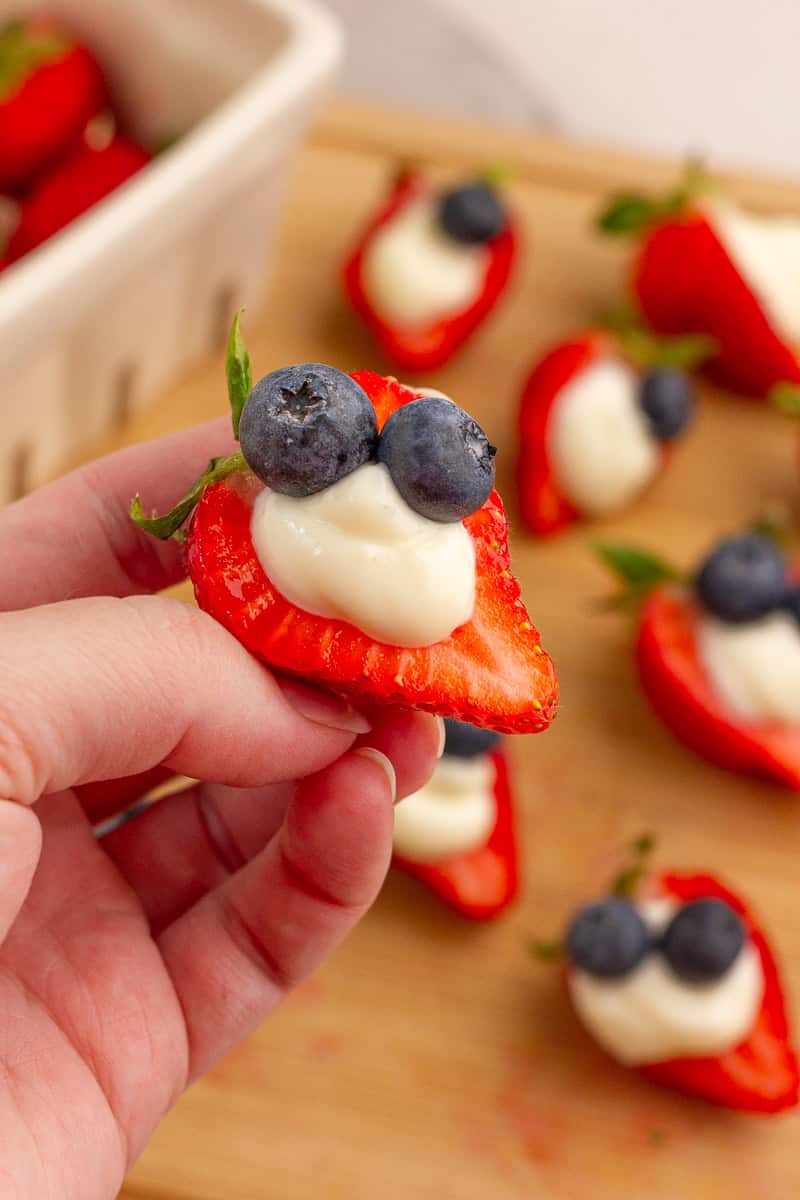 A hand holds a deviled strawberry filled with cheesecake filling and topped with blueberries.