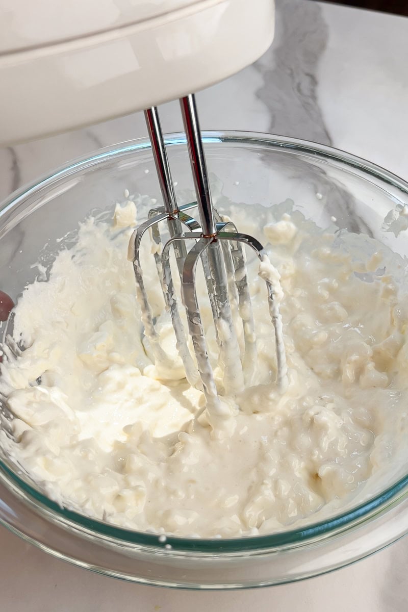 A hand mixer blends the cheesecake filling in a glass mixing bowl.