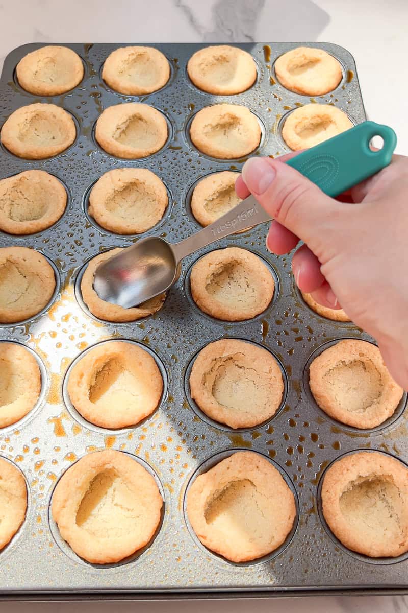A spoon presses warm sugar cookie cups into shape in a mini muffin pan.
