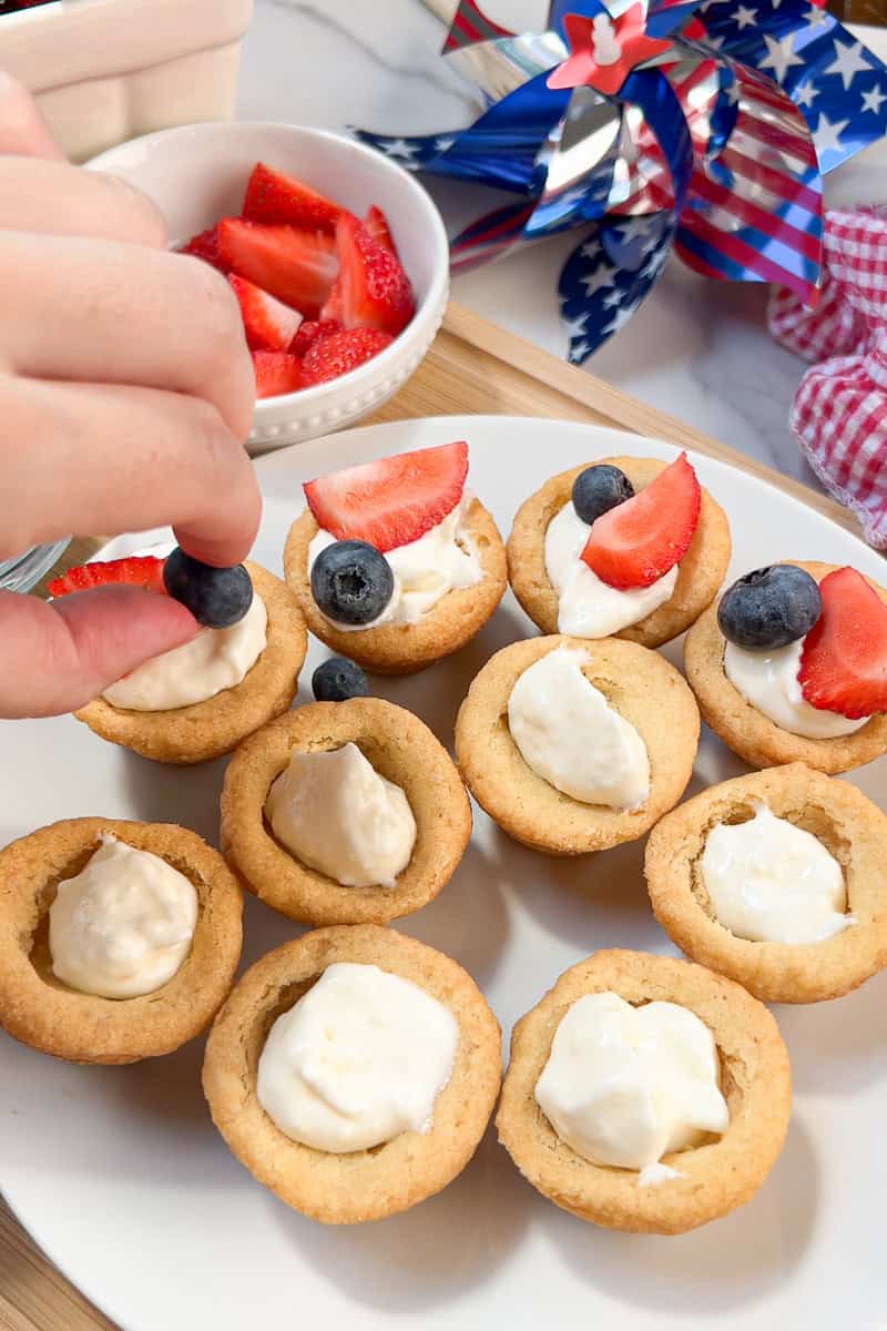 A hand tops fruit pizza cups with strawberries and blueberries on a white plate.