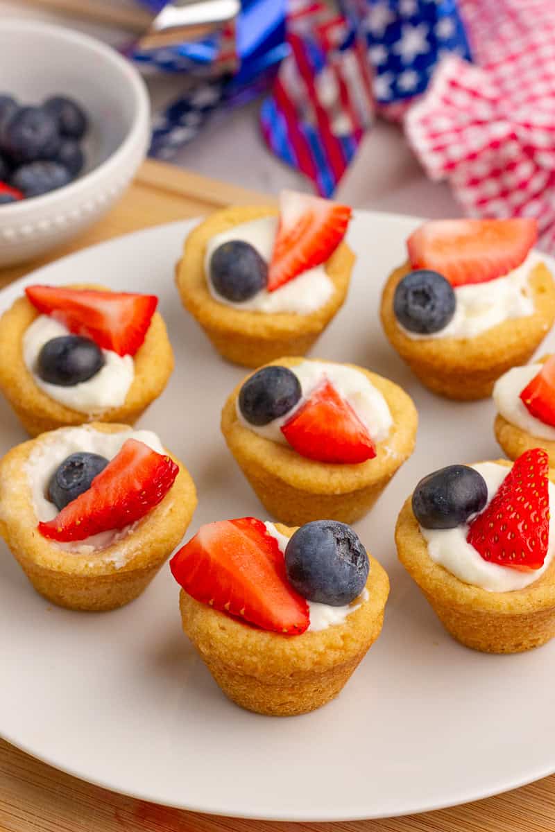 Finished fruit pizza cups sit on a white plate with fresh strawberries and blueberries.
