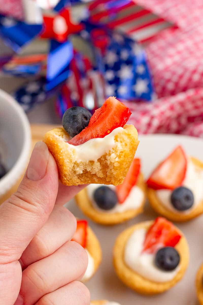 A hand holds a bite of a fruit pizza cup topped with strawberries and blueberries.