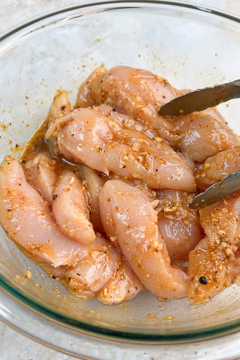 Raw chicken tenderloins are tossed in honey mustard marinade in a glass bowl.