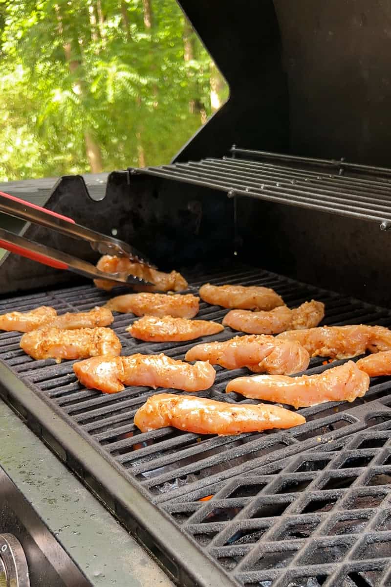 Marinated chicken tenderloins are placed on a hot grill with tongs.