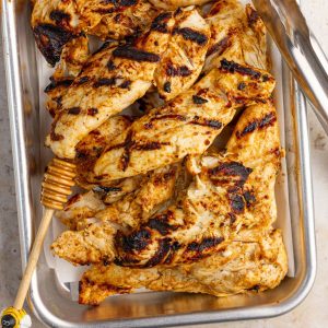 Grilled honey mustard chicken tenders are served in a tray with a honey dipper and tongs.