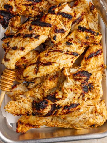 Grilled honey mustard chicken tenders are served in a tray with a honey dipper and tongs.