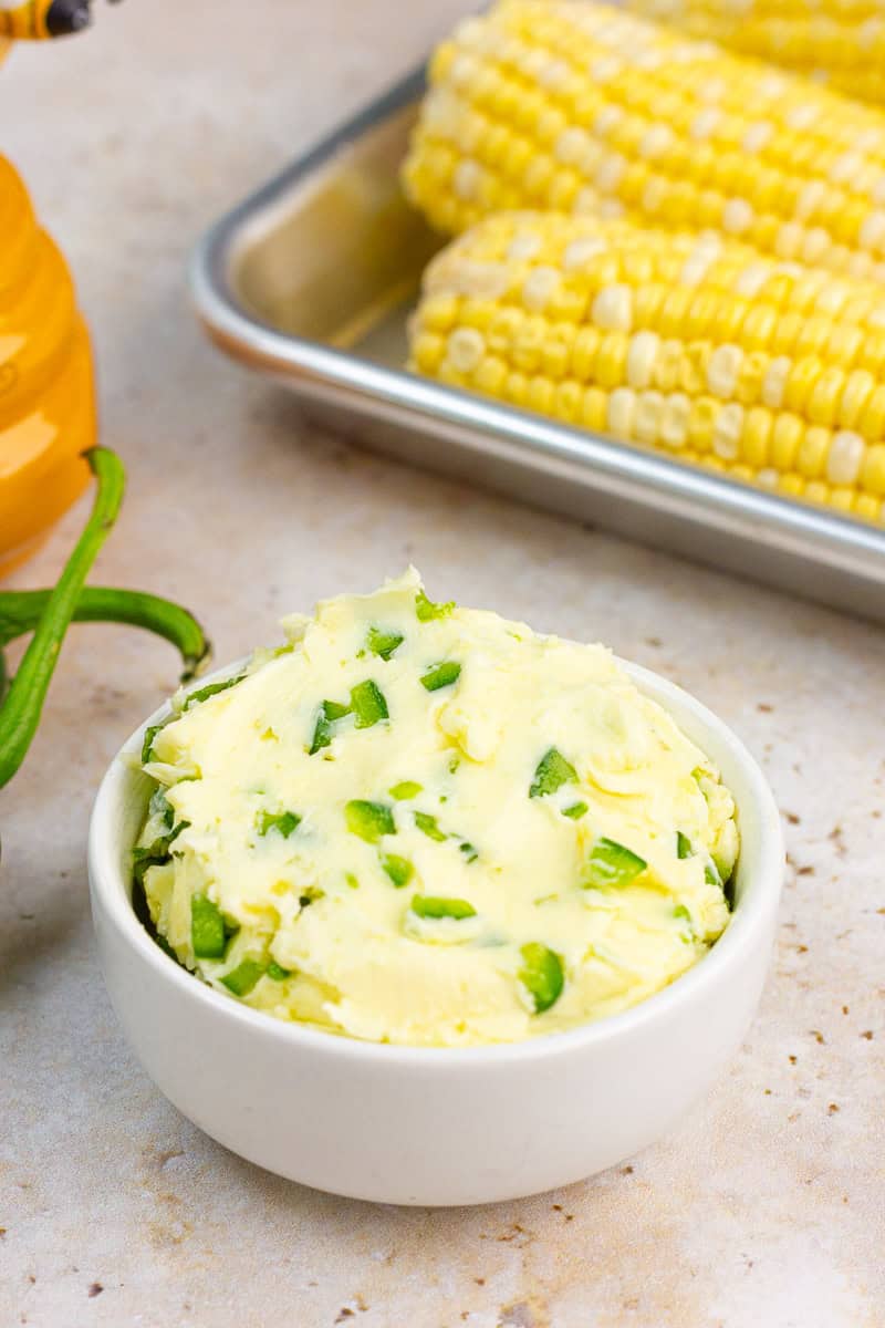 Jalapeno butter is served in a white bowl with corn on the cob and honey in the background.