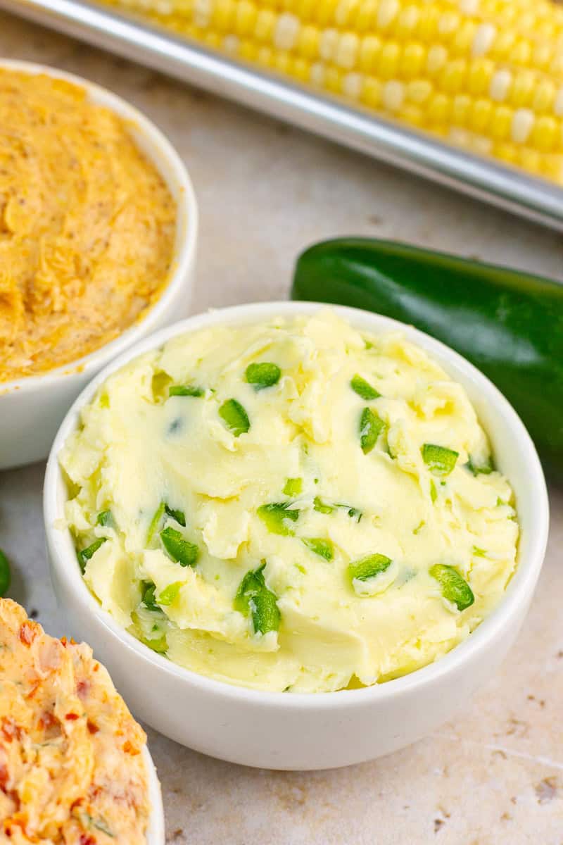 A bowl of jalapeno butter is served with corn on the cob, jalapeños, and various seasoned butter.