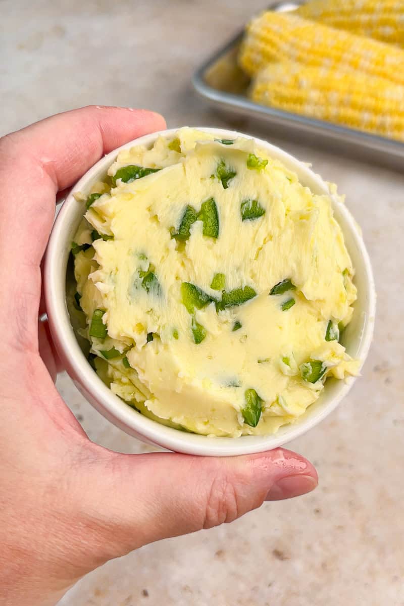 A hand holds a small bowl of creamy jalapeno butter with chopped jalapeños mixed throughout.