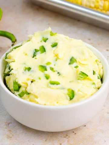Jalapeno butter is served in a white bowl with fresh jalapeño and corn on the cob in the background.