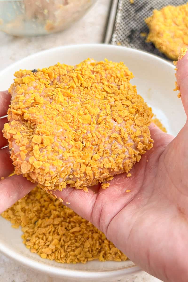 Hands pressing a ground chicken patty into crushed cornflake breading.
