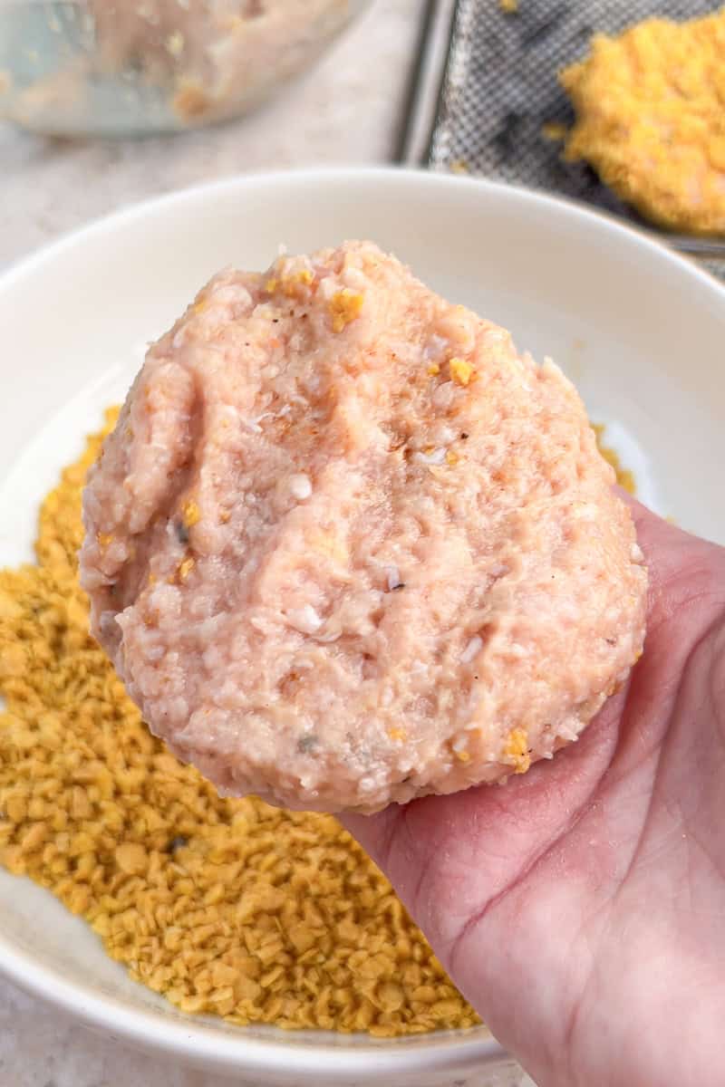 Hand shaping a ground chicken patty over a bowl of crushed cornflake breading.
