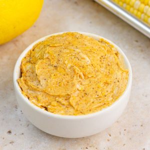 Creamy Old Bay butter is served in a white bowl with lemon and corn in the background.