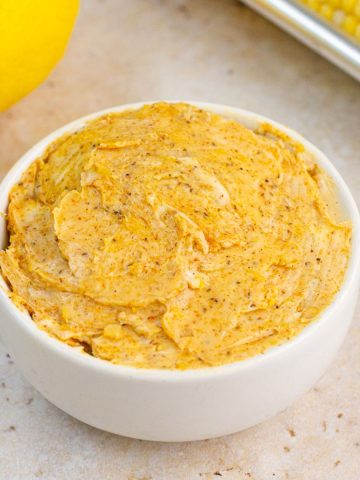 Creamy Old Bay butter is served in a white bowl with lemon and corn in the background.
