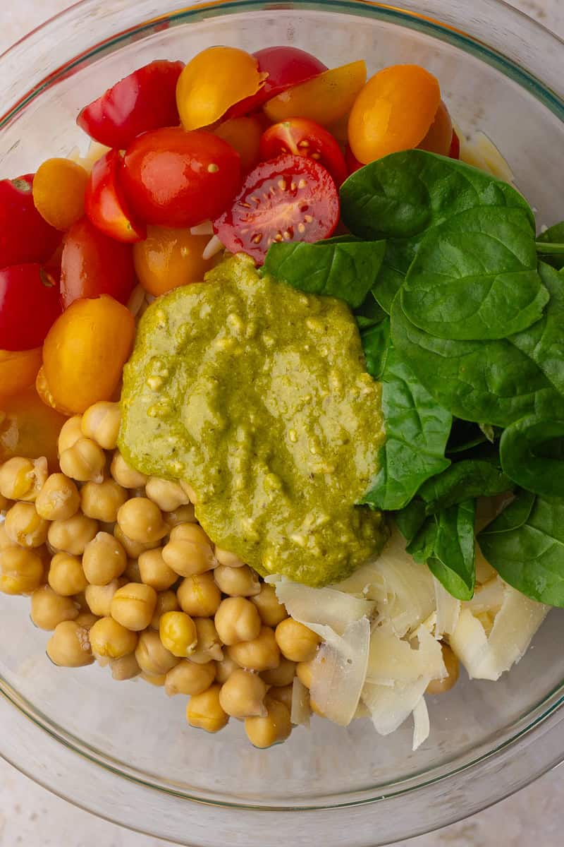 Orzo, chickpeas, spinach, tomatoes, and pesto are added to a bowl before mixing everything together.