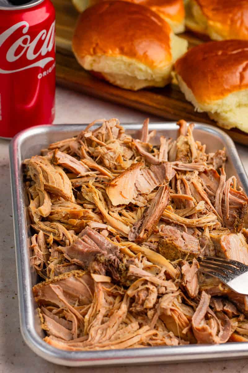 Tender shredded pulled pork sits in a metal pan with soft sandwich buns in the background.