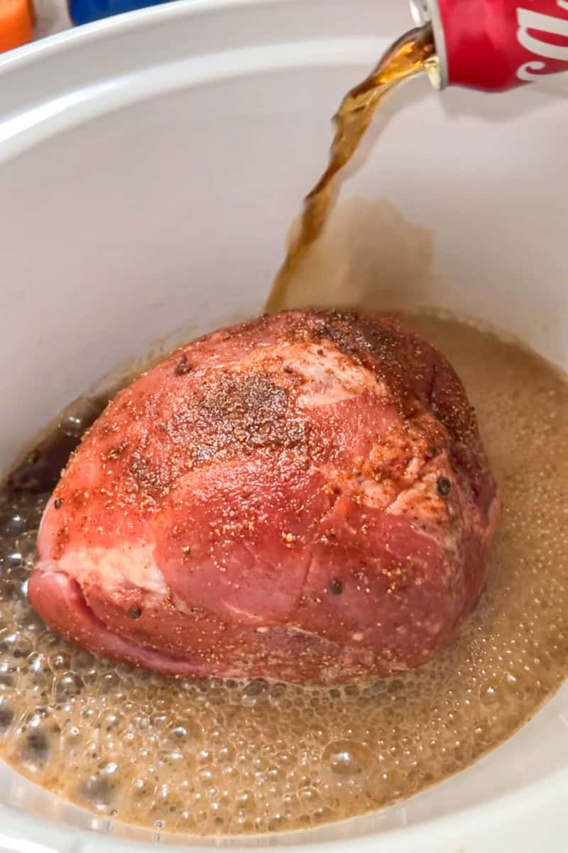 Coke is poured around seasoned pork shoulder in a slow cooker.
