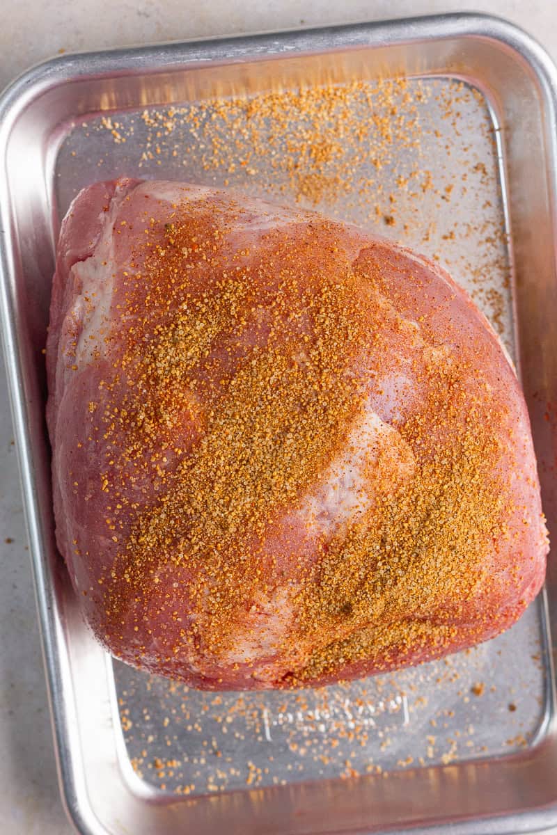 Raw pork shoulder is coated with BBQ dry rub seasoning in a metal pan.
