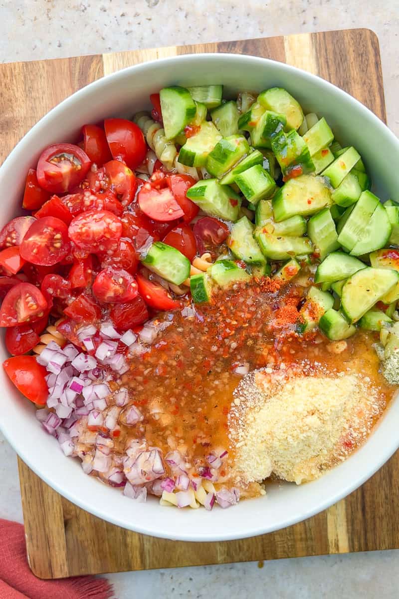 Pasta, cucumbers, tomatoes, red onion, parmesan, dressing, and seasoning are layered in a bowl.