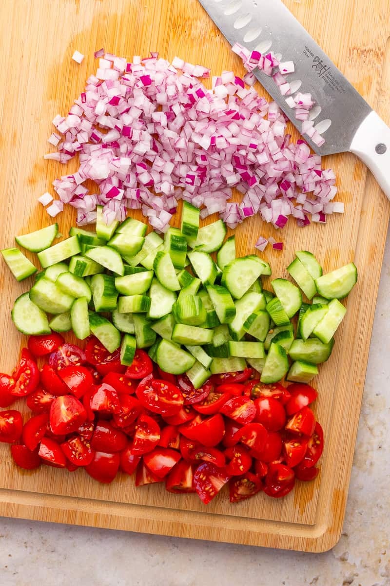 Chopped red onion, cucumbers, and cherry tomatoes sit on a cutting board for pasta salad.