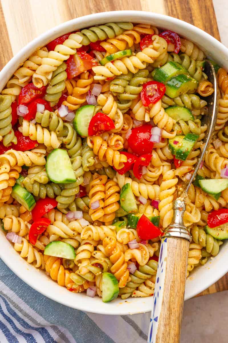 A close-up shows tri-color rotini pasta salad with cucumbers, cherry tomatoes, and red onion.