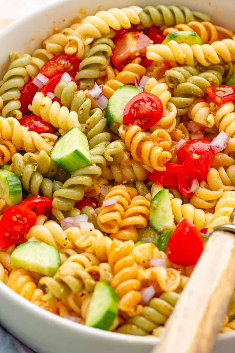 A close-up shows tri-color rotini pasta salad with cucumbers, cherry tomatoes, and red onion.