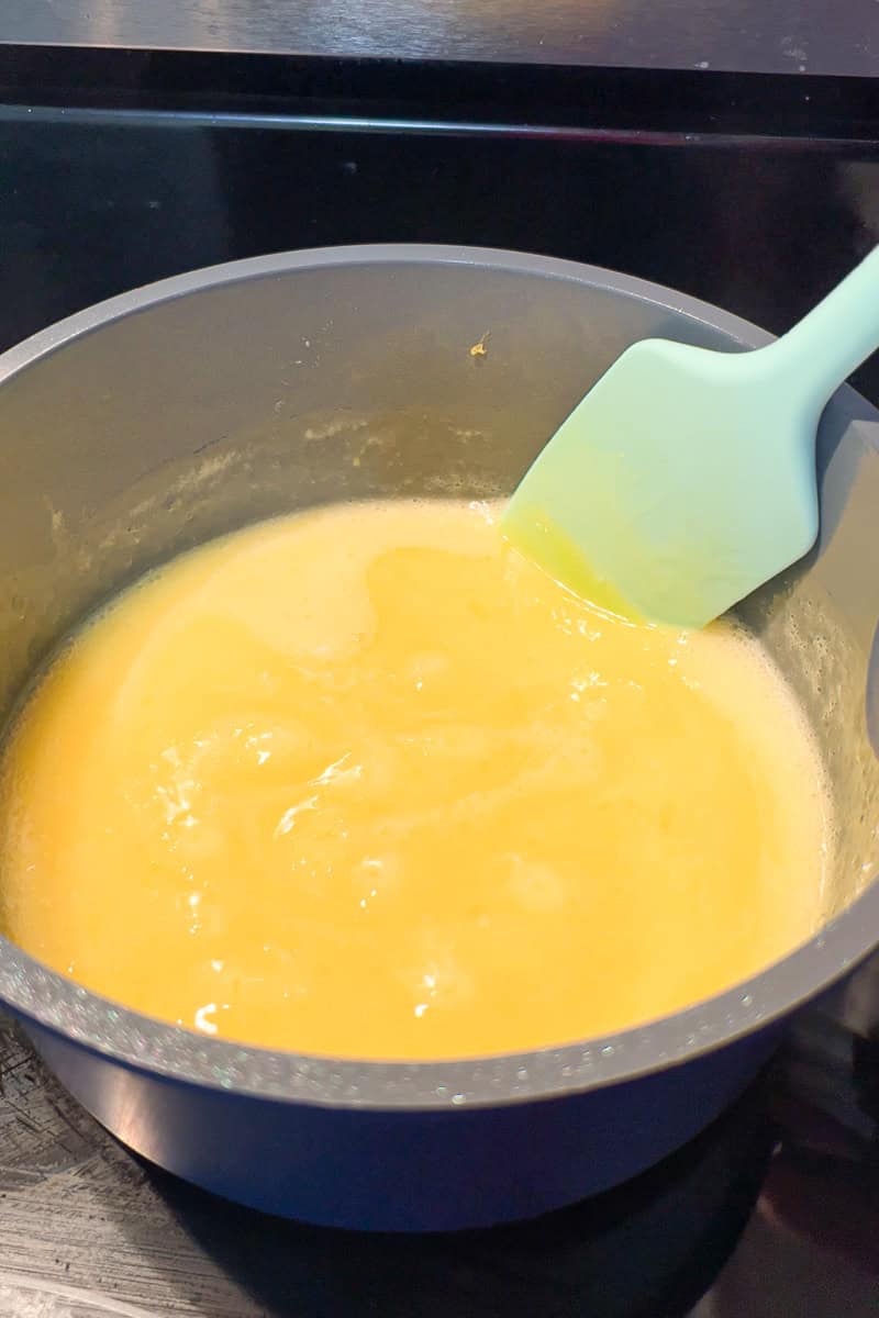 Homemade lemon curd cooks in a saucepan while a spatula stirs the smooth mixture.