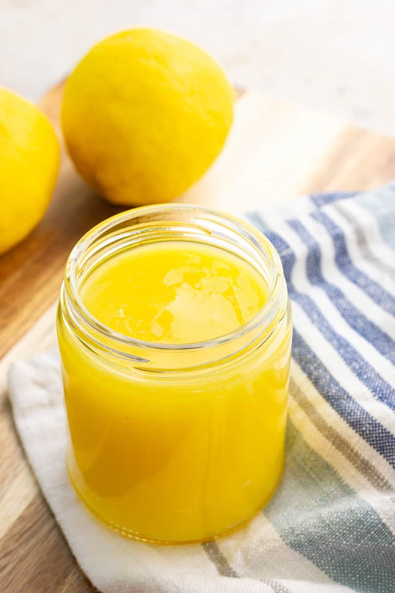 A jar of smooth small batch lemon curd sits on a kitchen towel beside fresh lemons.