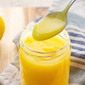 A spoonful of silky small batch lemon curd hovers over a glass jar filled with lemon curd.
