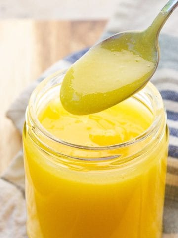 A spoonful of silky small batch lemon curd hovers over a glass jar filled with lemon curd.