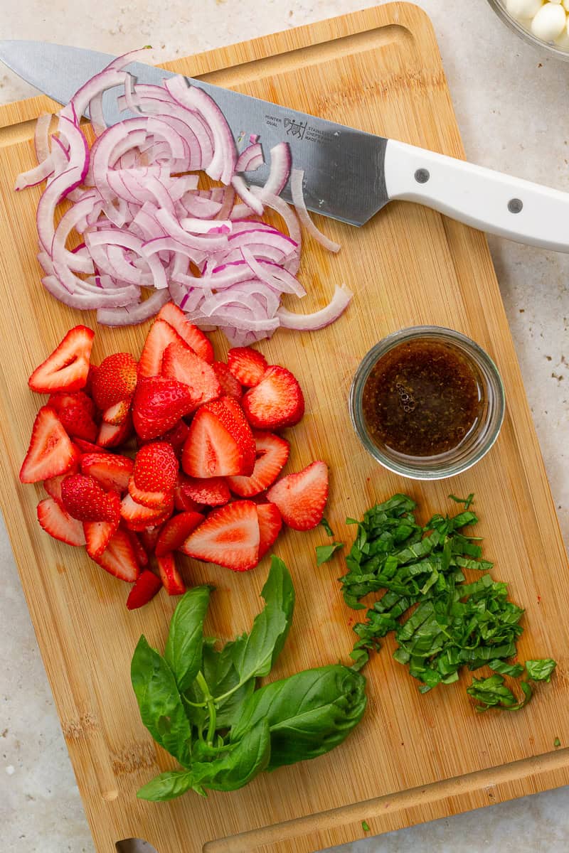 Sliced strawberries, red onion, fresh basil, and homemade balsamic dressing sit prepped on a cutting board for strawberry pasta salad.