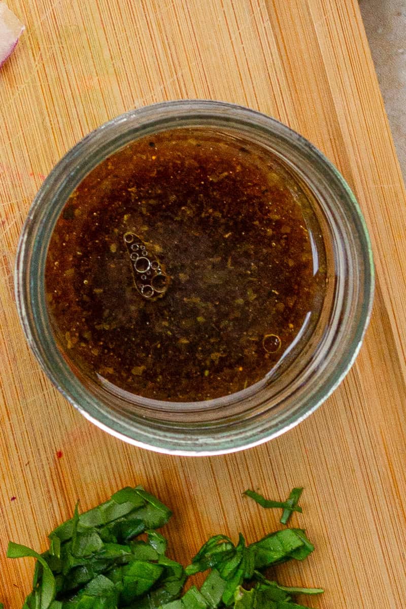 Homemade balsamic dressing for strawberry pasta salad sits in a small glass jar on a wooden cutting board.