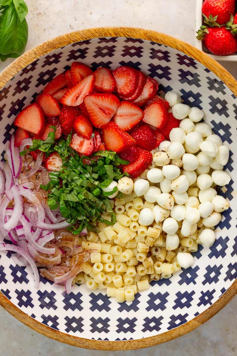 Cooked pasta, strawberries, mozzarella pearls, basil, red onion, and balsamic dressing are added to a large bowl before mixing.