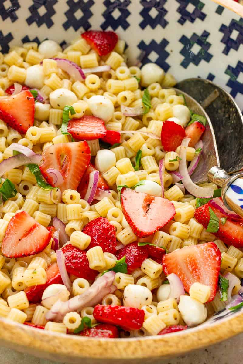 A close-up of strawberry pasta salad shows juicy strawberries, mozzarella pearls, basil, and red onion mixed into ditalini pasta.