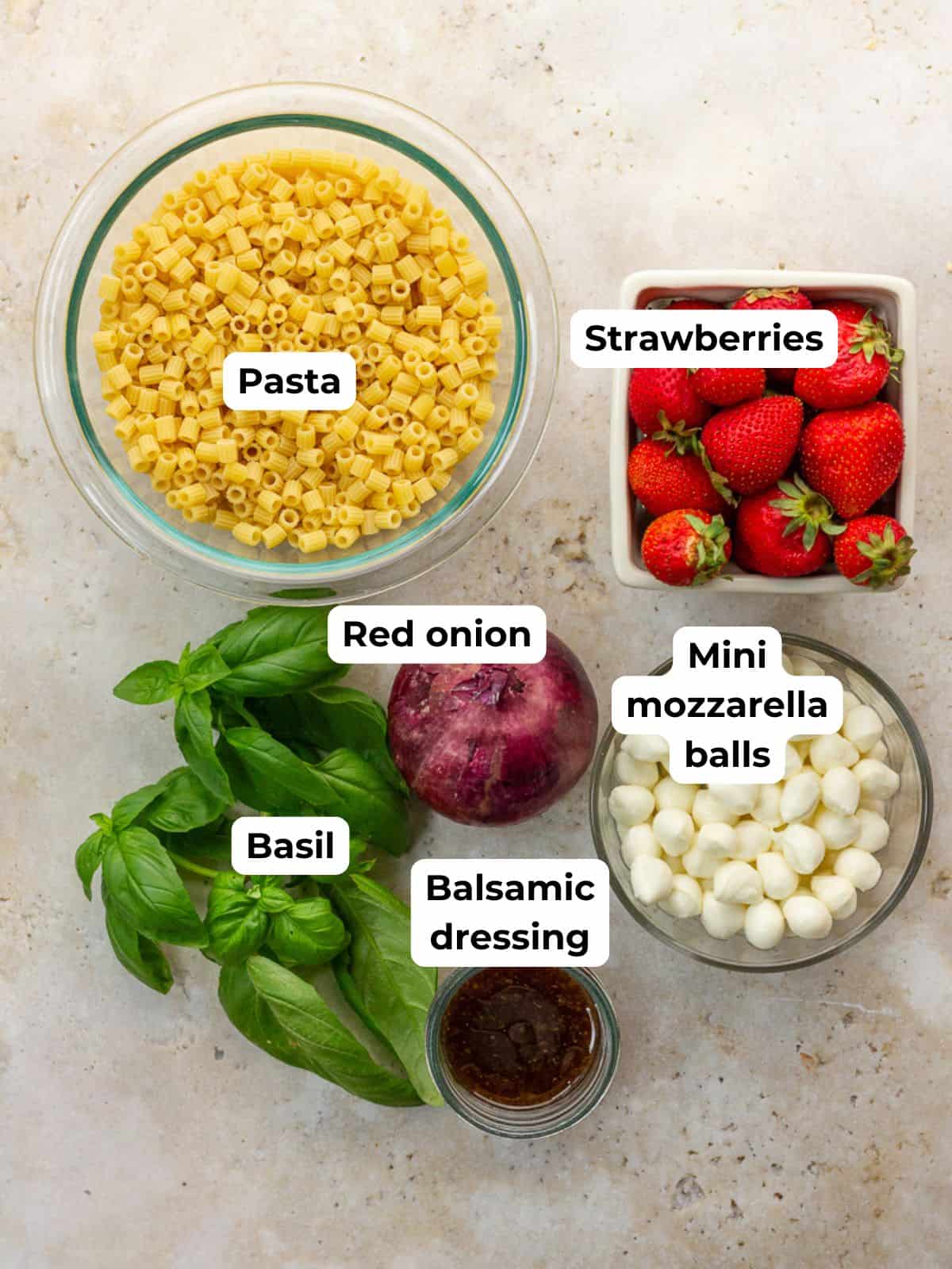 Ingredients for strawberry pasta salad including ditalini pasta, strawberries, mozzarella pearls, basil, red onion, and balsamic dressing are arranged on the counter.