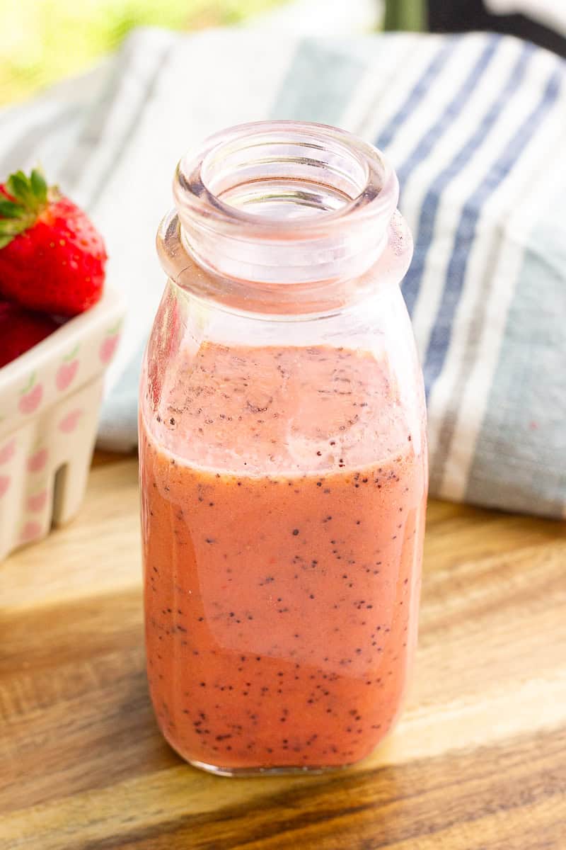 A jar of strawberry poppyseed dressing sits on a wooden board beside fresh strawberries.